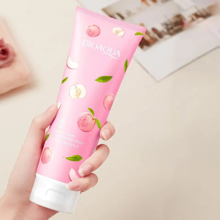 A pink tube of BIOAQUA Peach Extract Body Wash with sea salt, held in a hand, with a background that includes a photo and some decorative leaves.